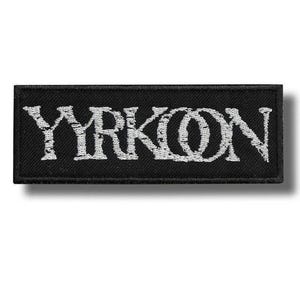 Op de afbeelding: Zwart rechthoekig embleem met het woord "YRKON" in witte, gestileerde letters geborduurd. Het embleem heeft een zwarte rand en is waarschijnlijk ontworpen om op kleding of accessoires te worden genaaid.