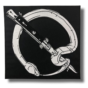 以下が含まれることがあります： 黒い正方形のパッチ。白い刺繍のデザインで、スプリングナイフに巻き付いたヘビが描かれています。ヘビは円を作り、ナイフがそれを貫通しています。パッチはクリーンでミニマリストな美学を持っています。