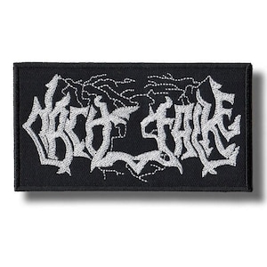May include: Black rectangular patch with white embroidered text that reads "DEATH FAKE" in a gothic font. The patch has a white border and is likely for clothing or accessories.