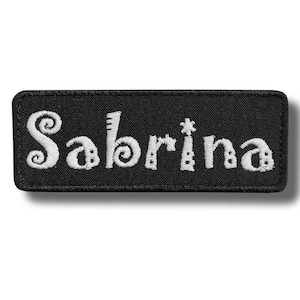 May include: Black rectangular patch embroidered with the name "Sabrina" in white. The letters are stylized with decorative swirls and dots. The patch has a black border and is likely designed to be sewn or attached to clothing or accessories.