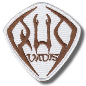 May include: Embroidered patch with a white background and a brown design. The design features stylized text and shapes within a diamond shape. The text "VADJS" is at the bottom of the patch.