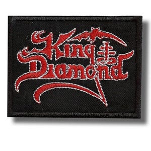 May include: Black rectangular patch with the words "King Diamond" in red, stylized lettering. The text is outlined in white stitching. A bat-like shape is above the word "King". The patch is likely for a jacket or bag.
