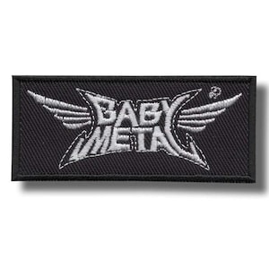 Puede incluir: Parche rectangular negro con las palabras "BABY METAL" en blanco, con letras estilizadas. El diseño incluye alas a ambos lados del texto. El parche es probablemente para ropa o accesorios.