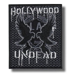 Puede incluir: Parche bordado en negro y gris con las palabras "HOLLYWOOD UNDEAD" y un diseño de escudo con dos aves estilizadas. El parche tiene un fondo texturizado y detalles blancos goteando.