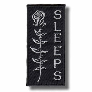 Könnte beinhalten: Schwarzer rechteckiger Aufnäher mit einer weißen, gestickten Rose und einem Stiel. Das Wort "SLEEPS" ist vertikal in weißen Großbuchstaben auf der rechten Seite gestickt. Der Aufnäher hat einen schwarzen Rand.