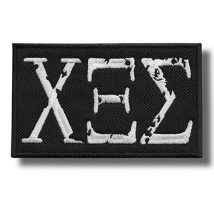Peut inclure: Écusson rectangulaire noir avec les lettres grecques X, E et Σ brodées en blanc. Les lettres présentent un aspect texturé et vieilli. Idéal pour les vêtements ou accessoires.