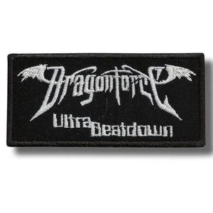 Pode incluir: Patch retangular preto com texto bordado branco. O texto diz "Dragonforce" em uma fonte estilizada, com "Ultra Beatdown" abaixo. O patch tem uma borda preta e um design de asa branca em cada lado do texto principal.