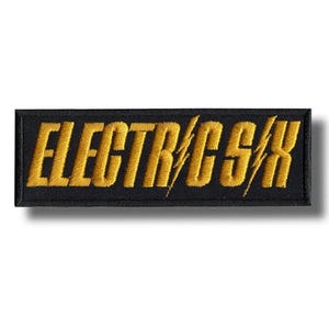 Puede incluir: Parche rectangular negro con el texto bordado en oro "ELECTRIC CSX". Un diseño de rayo está incorporado en el texto. El parche tiene un borde negro y es probablemente un emblema o logotipo.
