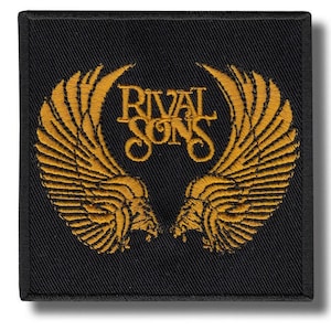May include: Black square patch with gold embroidered design. The words "RIVAL SONS" are in gold, above a pair of symmetrical gold wings. The patch has a textured surface, with the gold elements contrasting against the black.