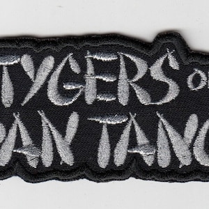 Peut inclure: Écusson brodé noir avec l'inscription "TYGERS of PAN TANG" en fil argenté. Le patch a une bordure noire et une forme irrégulière, idéal pour personnaliser des vêtements ou des accessoires.