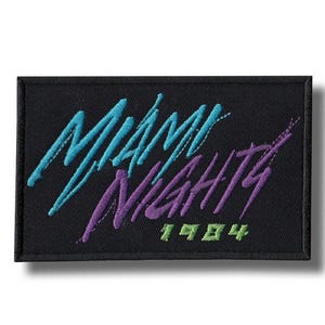 Op de afbeelding: Zwart rechthoekig embleem met de woorden "Miami Nights" in turquoise en paars schrift. Het jaar "1984" is eronder in groen geborduurd. De patch heeft een zwarte rand.