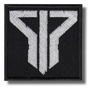 Op de afbeelding: Zwart vierkantige patch met een wit geborduurd geometrisch ontwerp. Het ontwerp heeft een gestileerde letter "T" met een gedurfde, hoekige esthetiek. De patch heeft een zwarte rand en een getextureerde uitstraling.