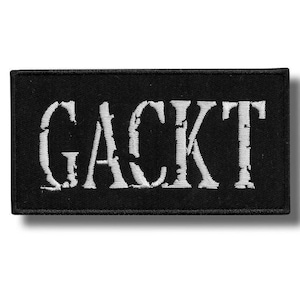 Pode incluir: Patch retangular preto com o texto branco "GACKT". O texto tem uma aparência desgastada e texturizada. O patch é provavelmente bordado ou impresso, possivelmente para roupas ou acessórios.