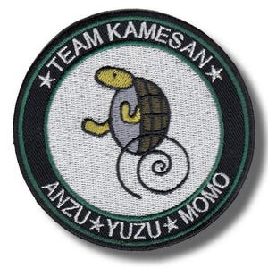 Könnte beinhalten: Runder, gestickter Aufnäher mit einem Cartoon-Schildkröten-Design. Der Aufnäher trägt den Text "TEAM KAMESAN" und "ANZU * YUZU * MOMO" in weißen Buchstaben. Die Schildkröte ist grau, braun und gelb, mit einem spiralförmigen Schwanz. Der Aufnäher hat einen schwarzen und grünen Rand.