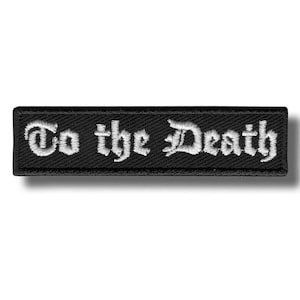 Puede incluir: Parche rectangular negro con la frase bordada "To the Death" en letras góticas blancas. El parche tiene un borde negro y está sobre un fondo blanco.
