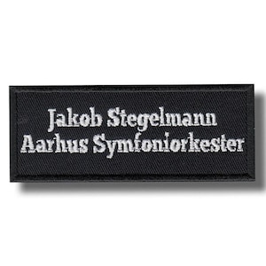 Puede incluir: Una placa rectangular negra con texto bordado en blanco. El texto dice "Jakob Stegelmann Aarhus Symfoniorkester". Esta placa es probablemente para un miembro de la Orquesta Sinfónica de Aarhus.