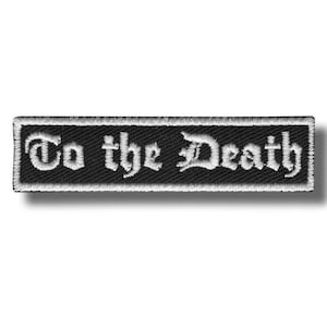 以下が含まれることがあります： 白いゴシック体で「To the Death」と書かれた黒い刺繍パッチ。パッチは白い縁取りがあり、長方形です。衣類やアクセサリーに最適です。