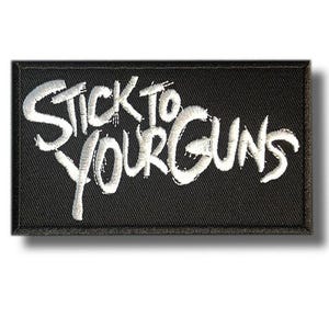 Può includere: Toppa rettangolare nera con testo ricamato bianco « STICK TO YOUR GUNS ». La toppa ha un bordo nero e un effetto ombra. La scritta è in un carattere corsivo stilizzato, dando un aspetto strutturato.