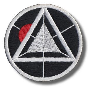 May include: Round black patch with a white embroidered geometric design. The design includes a triangle, lines, and a red circle. The patch has a white border and is likely an emblem or insignia.