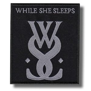 May include: Black square patch with the text "WHILE SHE SLEEPS" embroidered in white above a stylized gray logo. The logo is a combination of the letters "W" and "S" intertwined.