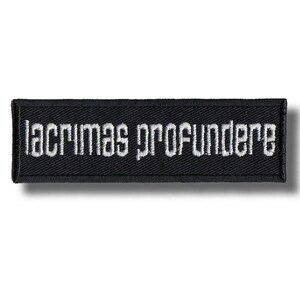 Könnte beinhalten: Ein rechteckiger, schwarzer Aufnäher mit dem weißen, gestickten Text "lacrimas profundere". Der Aufnäher hat einen schwarzen Rand und ist zum Aufnähen oder Aufbügeln auf Stoff gedacht. Der Text ist in einer stilisierten Schriftart.