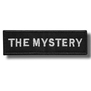 Könnte beinhalten: Ein rechteckiger schwarzer Aufnäher mit dem gestickten Text "THE MYSTERY" in Weiß. Der Aufnäher hat einen schwarzen Rand und ist zum Aufnähen oder Aufbügeln auf Kleidung oder Accessoires gedacht.
