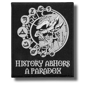 May include: Black embroidered patch featuring a detailed white illustration of a stylized face within a circular design. The text "HISTORY ABHORS A PARADOX" is embroidered in white below the image. The patch has a rectangular shape.