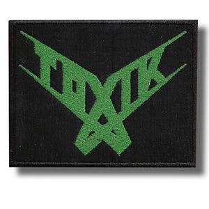 Pode incluir: Patch quadrado preto com um design de texto verde estilizado. O texto verde parece ser uma palavra ou símbolo abstrato. O patch tem uma borda preta e é provavelmente feito de tecido.