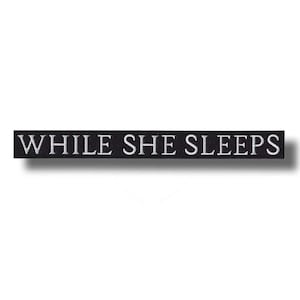 Könnte beinhalten: Schwarzer rechteckiger Aufnäher mit dem gestickten weißen Text "WHILE SHE SLEEPS". Der Aufnäher hat eine lange, schmale Form, und der Text ist zentriert. Der Aufnäher ist wahrscheinlich zum Aufnähen auf Kleidung oder Accessoires gedacht.