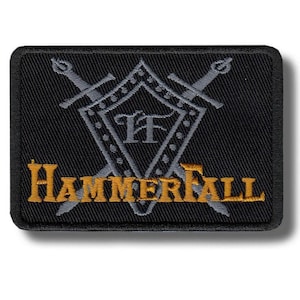 Może przedstawiać: Czarna prostokątna naszywka z wyhaftowanym złotym napisem "HammerFall". Naszywka posiada szary wzór tarczy z dwoma skrzyżowanymi mieczami i literami "TF" pośrodku. Naszywka ma czarną obwódkę.