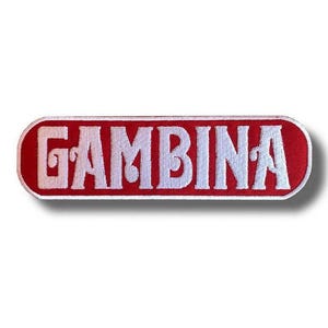 Puede incluir: Un parche rectangular rojo con esquinas redondeadas, con la palabra "GAMBINA" bordada en blanco. El parche está diseñado para su aplicación en ropa o accesorios.