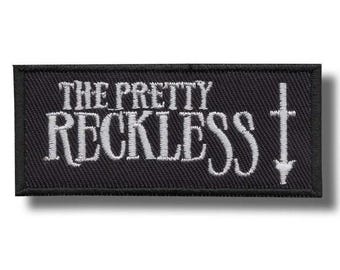 May include: A rectangular black patch with white embroidered text that reads "THE PRETTY RECKLESS". A white cross with a downward arrow is on the right side. The patch is likely for clothing or accessories.