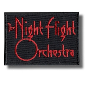 May include: A black rectangular patch embroidered with the words "The Night Flight Orchestra" in red. The text is in a stylized font, with "Night" arched above "Orchestra". The patch has a black border and a shadow.
