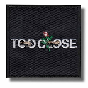 Peut inclure: Écusson carré noir avec l'inscription brodée en blanc "TOO CLOSE" et un motif de rose. La rose est rouge avec des feuilles vertes et une tige marron. L'écusson a une bordure noire, idéal pour coudre ou repasser sur du tissu.