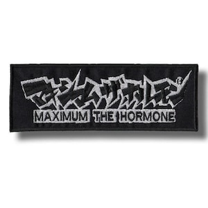 以下が含まれることがあります： 黒い長方形のパッチ。白い刺繍で「MAXIMUM THE HORMONE」と書かれています。パッチは白い縁取りと影があります。テキストは様式化されたフォントです。
