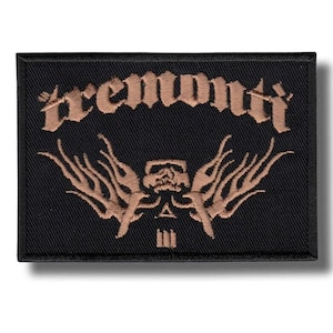 Op de afbeelding: Zwart rechthoekig embleem met het woord "Tremonti" in een gestileerd lettertype boven een bronzen ontwerp met een schedel, vlammen en geometrische vormen.