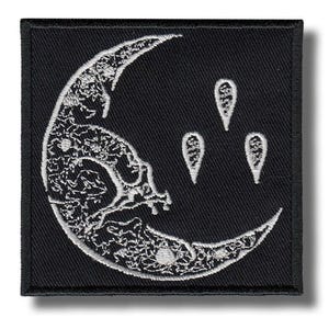 May include: A square black patch featuring a white embroidered crescent moon design, adorned with floral patterns and three teardrop shapes. The patch has a black border, suitable for clothing or accessories.