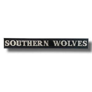 May include: A black rectangular sign with the words "SOUTHERN WOLVES" in a decorative, off-white font. The sign is likely made of wood or a similar material, and the text is centered.