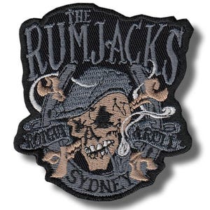 Puede incluir: Parche bordado con una calavera con sombrero de pirata, espadas cruzadas y el texto "THE RUMJACKS SYDNEY". El parche es principalmente negro, con detalles grises, beige y blancos. El diseño tiene un aspecto vintage.