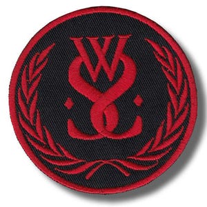 May include: Circular black patch with a red embroidered design. The design features intertwined letters and a surrounding laurel wreath. The patch has a red border and is likely for clothing or accessories.