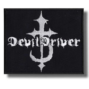 Puede incluir: Parche rectangular negro con texto bordado blanco y un diseño de cruz estilizada. El texto "DevilDriver" está escrito en una fuente gótica, con la letra "D" incorporada en la cruz. El parche tiene un borde negro.
