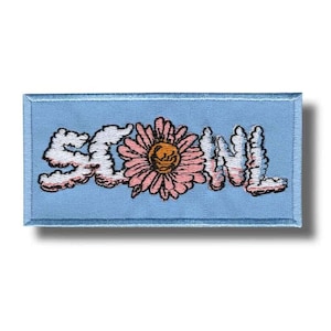 Pode incluir: Um patch bordado azul claro com a palavra "SLOW" em letras brancas em forma de nuvem e uma margarida rosa. A margarida tem um centro amarelo. O patch é retangular.