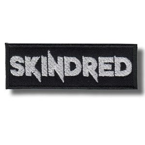 Könnte beinhalten: Ein rechteckiger schwarzer Aufnäher mit dem Wort "SKINDRED" in weiß gestickten, stilisierten Buchstaben. Der Aufnäher hat einen schwarzen Rand und ist wahrscheinlich zum Aufnähen auf Kleidung oder Accessoires gedacht.