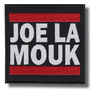 Può includere: Toppa quadrata nera con la scritta bianca "JOE LA MOUK". Il testo è centrato sulla toppa, con una striscia orizzontale rossa sopra e sotto il testo. La toppa ha un bordo nero.