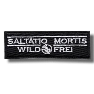 Peut inclure: Écusson rectangulaire noir avec texte brodé blanc. Le texte indique "SALTATIO MORTIS" au-dessus de "WILD" et "FREI", avec un emblème circulaire au centre. L'écusson est probablement un accessoire, pour vêtements ou sacs.