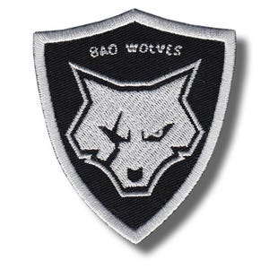 May include: A black and white embroidered patch in a shield shape. The patch features the text "BAD WOLVES" at the top and a stylized wolf head in the center. The patch has a white border and is likely for clothing or accessories.