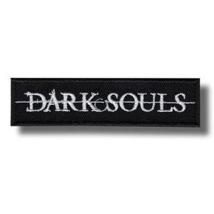 Könnte beinhalten: Ein rechteckiger, schwarzer Aufnäher mit dem weißen Schriftzug "DARK SOULS". Der Text ist zentriert und in einer stilisierten Schriftart gehalten. Der Aufnäher ist wahrscheinlich für Kleidung oder Accessoires gedacht.