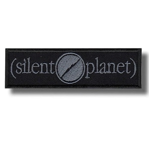 May include: Black rectangular patch with the text "(silent planet)" embroidered in light gray. A gray circle with a diagonal line is in the center. The patch is likely for clothing or accessories.