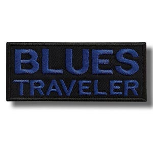 May include: A rectangular black patch with the words "BLUES TRAVELER" embroidered in blue. The text is in a bold, sans-serif font, and the patch has a black border.
