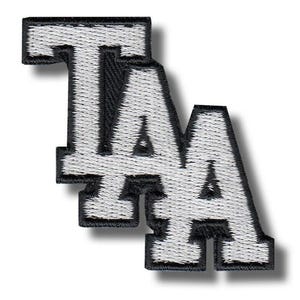 May include: Embroidered patch displaying the letters "TA" stacked. The letters are white with black outlining and a black border. The patch has a textured appearance, likely made from fabric or thread. The background is plain white.